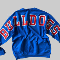 A bright blue long-sleeve sweatshirt laid flat on a beige fabric ottoman, showing the back with large red and white 'BULLDOGS' lettering across the upper back.