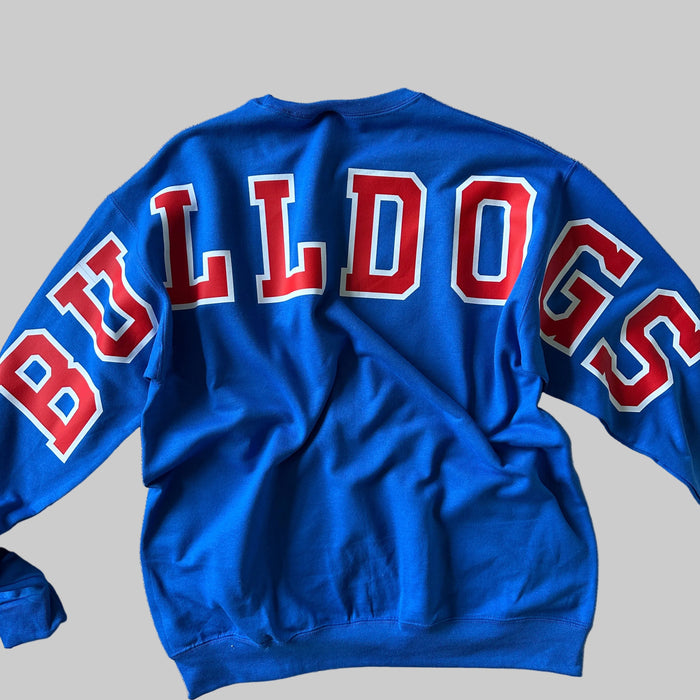 A bright blue long-sleeve sweatshirt laid flat on a beige fabric ottoman, showing the back with large red and white 'BULLDOGS' lettering across the upper back.
